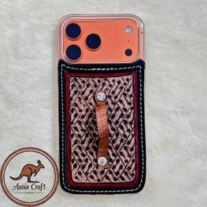 Puede incluir: Una funda transparente para teléfono con una cartera de cuero estampada adjunta. La cartera es de color canela con un diseño negro y una correa de cuero marrón con dos gemas plateadas. El teléfono es naranja.