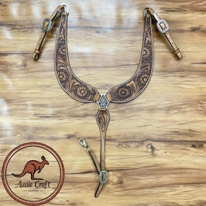 Op de afbeelding: Een lichtbruine leren borsttuig voor paarden met bloemmotieven. De borsttuig heeft zilveren gespen en accenten. De afbeelding bevat ook een logo met een kangoeroe en de tekst "Aussie Craft Leather".