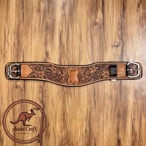 May include: Aussie Craft leather horse cinch. The brown cinch with a black border features a floral design. It has silver buckles and D-rings. The cinch is displayed on a wooden background.