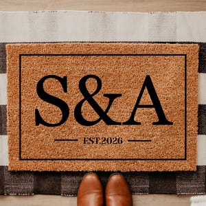 May include: A rectangular doormat with a brown coir surface, featuring the black letters "S&A" above "EST.2026". The mat is framed with a black border and rests on a black and white checkered rug. Brown shoes are visible at the bottom.