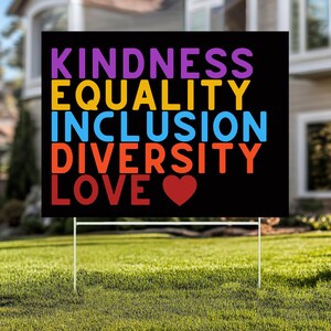 May include: A black yard sign with the words "KINDNESS, EQUALITY, INCLUSION, DIVERSITY, LOVE" in purple, yellow, blue, orange, and red. A red heart graphic is below the word "LOVE". The sign is on a white metal stand, placed in a grassy yard.
