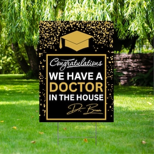 May include: A black and gold graduation yard sign with the text "Congratulations WE HAVE A DOCTOR IN THE HOUSE" and a graduation cap graphic. The sign is displayed on a metal stake in a grassy yard.