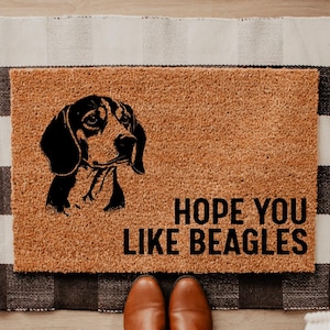 May include: A rectangular doormat made of natural coir fiber, featuring a black and white beagle illustration and the text "HOPE YOU LIKE BEAGLES". The mat is placed on a black and white checkered rug.