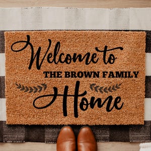 May include: A rectangular tan coir doormat featuring black text that reads Welcome to THE BROWN FAMILY Home, accented with two small black leaf sprigs. The mat has a coarse, natural fiber texture and is centered on a black and white plaid rug.