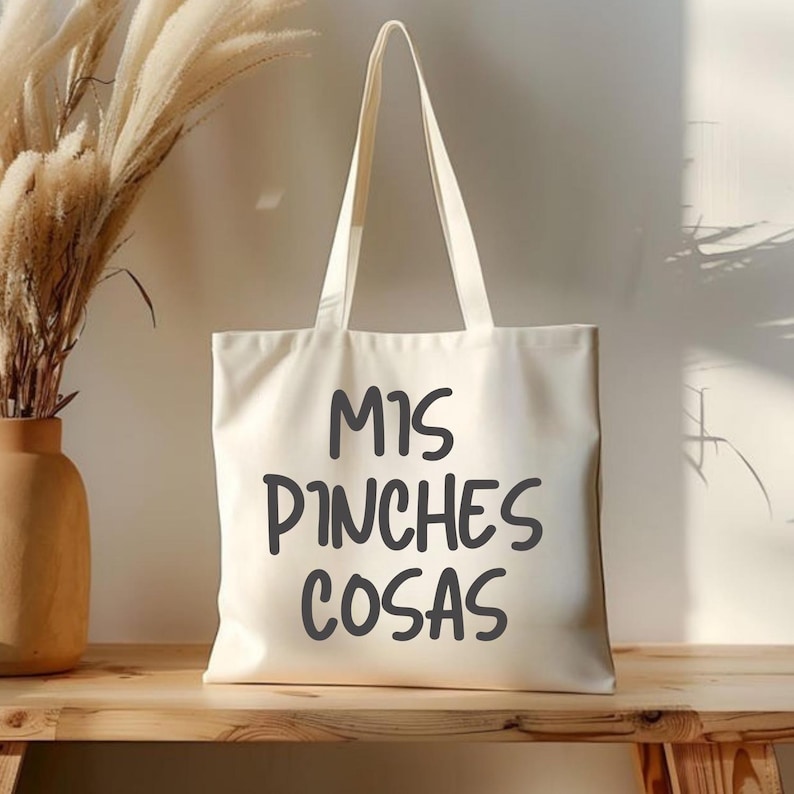 Puede incluir: Bolso tote de color crema con el texto "MIS PINCHES COSAS" en gris oscuro. El bolso tiene dos asas y est&aacute; hecho de una tela resistente. El bolso tote est&aacute; sobre una superficie de madera.