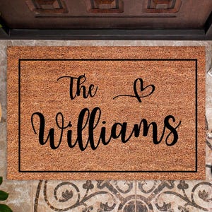 May include: A rectangular doormat made of natural coir fiber, featuring the words "The Williams" in elegant black script with a heart symbol. The mat is framed with a black border and is placed in front of a door.