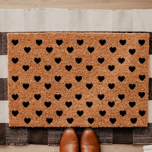 May include: A rectangular doormat with a tan background and a pattern of black hearts. The mat is placed on top of a black and white checkered rug. Brown leather shoes are visible at the bottom of the image.