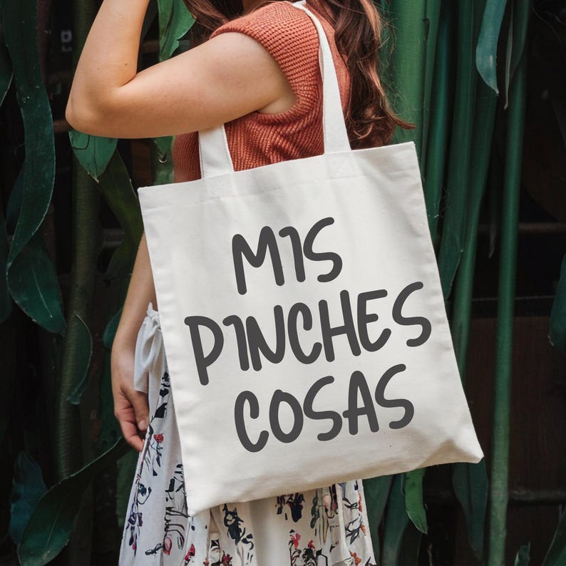 Puede incluir: Bolso tote de color crema con la frase "MIS PINCHES COSAS" impresa en gris oscuro. El bolso tiene dos asas blancas y se muestra sobre un fondo de hojas verdes.
