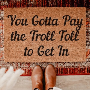 May include: A rectangular doormat made of natural fibers, featuring the phrase "You Gotta Pay the Troll Toll to Get In" in black script. The mat is placed on a patterned rug with brown leather boots in the foreground.