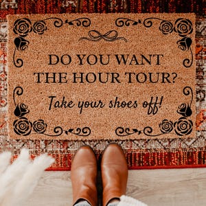 May include: A rectangular doormat with the text "DO YOU WANT THE HOUR TOUR? Take your shoes off!" in black. The mat is brown with a decorative black floral border. Brown boots are visible at the bottom of the image.
