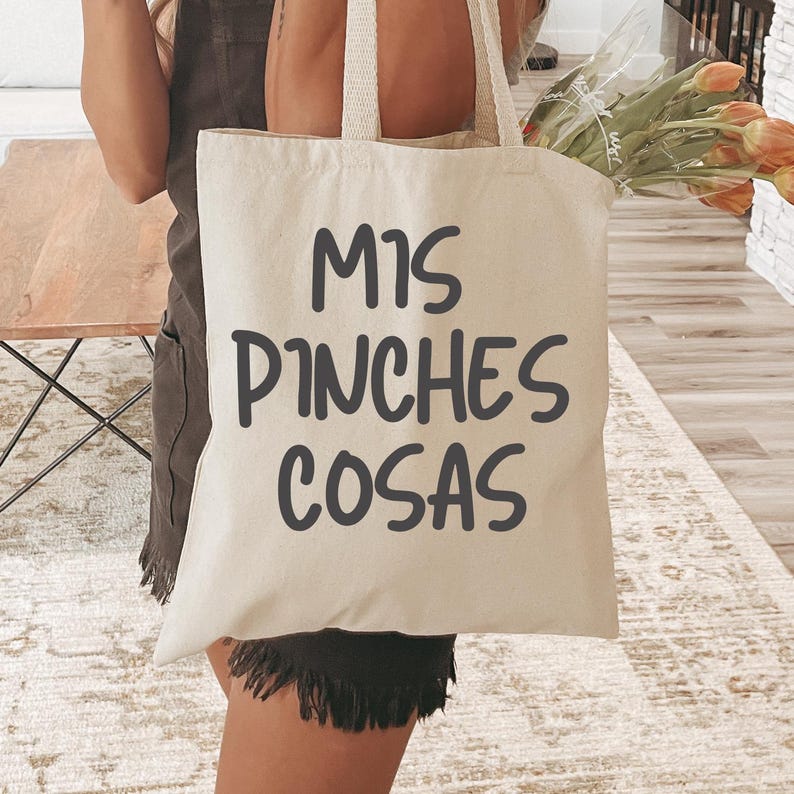 Puede incluir: Un bolso tote de lona beige con el texto "MIS PINCHES COSAS" en letras de color gris oscuro. El bolso tiene un dise&ntilde;o sencillo con dos asas y lo sostiene una persona. Se ve un ramo de tulipanes naranjas.