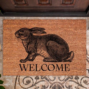 May include: A rectangular doormat made of natural coir fiber. The mat features a detailed illustration of a rabbit in dark brown ink, with the word "WELCOME" printed in black below. The mat is placed in front of a wooden door.