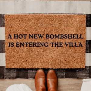 May include: A rectangular doormat with the text "A HOT NEW BOMBSHELL IS ENTERING THE VILLA" in black. The mat is brown and sits on a black and white checkered rug. Brown leather boots are visible.