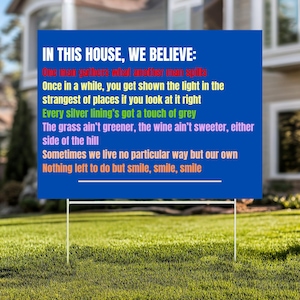 May include: A blue rectangular sign with white text on a metal frame, displayed on a grassy lawn. The sign reads, "IN THIS HOUSE, WE BELIEVE:" followed by a list of inspirational quotes. The sign is outdoors, in front of a house.