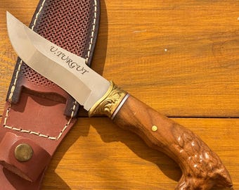 May include: A handcrafted hunting knife with a polished steel blade and a wooden handle. The knife features a brass bolster and is accompanied by a brown leather sheath. The blade is engraved with the text "U.TURGUT".