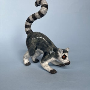 May include: A ceramic lemur figurine featuring a glossy crackle glaze finish in shades of grey, white, and black. The lemur is depicted in a crouching stance with its long, striped tail curved upward, and it has distinct orange-coloured eyes.