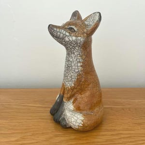 May include: A ceramic fox ornament with a crackle glaze. The fox is depicted in a seated position, with its head turned. The fur is a blend of brown, white, and black. The ornament is displayed on a wooden surface.
