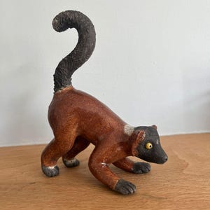 May include: A ceramic figurine of a lemur with a glossy reddish-brown body, a dark textured tail curved upwards, and a black face with a single visible yellow eye. The animal is depicted in a low, crouching stance with its front limbs extended.