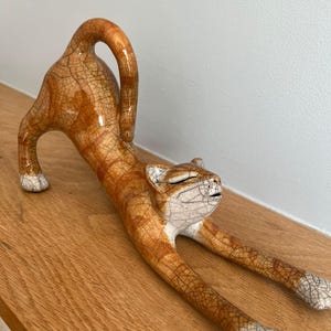 May include: A ceramic cat figurine in a stretching pose, with orange and white coloring and a cracked glaze finish. The cat is lying on a wooden surface.
