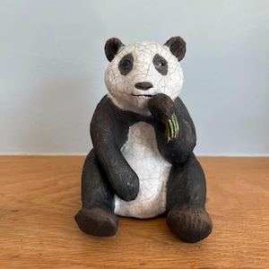 May include: A ceramic panda figurine in a seated position, featuring a white face and belly with a distinct black crackle glaze finish. The panda has dark brown limbs and ears, and is holding a small, green-tinted bamboo shoot to its mouth.