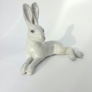May include: A ceramic hare figurine in a reclining position with long, upright ears. The surface features a white glaze with a distinct dark grey crackle pattern throughout the body, head, and ears, giving it a textured, weathered appearance.