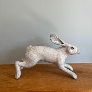 May include: A ceramic hare figurine in a dynamic running pose, featuring a white base colour with a prominent black crackle glaze finish. The sculpture displays elongated ears, a small tail, and a smooth, glossy surface texture throughout.