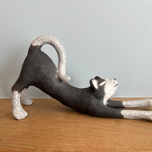 May include: A ceramic cat figurine in a stretching pose. The cat is primarily dark gray with white accents on the paws, ears, and tail. The tail is arched, and the cat's eyes are closed. The figurine is on a wooden surface.