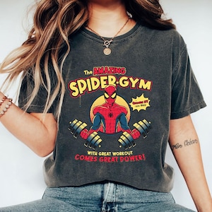 Może przedstawiać: Ciemnoszara koszulka z grafiką Spider-Mana podnoszącego ciężary, z napisem "The Amazing Spider-Gym". Projekt zawiera frazę "With great workout comes great power!" oraz ż&oacute;łto-czerwoną kolorystykę.