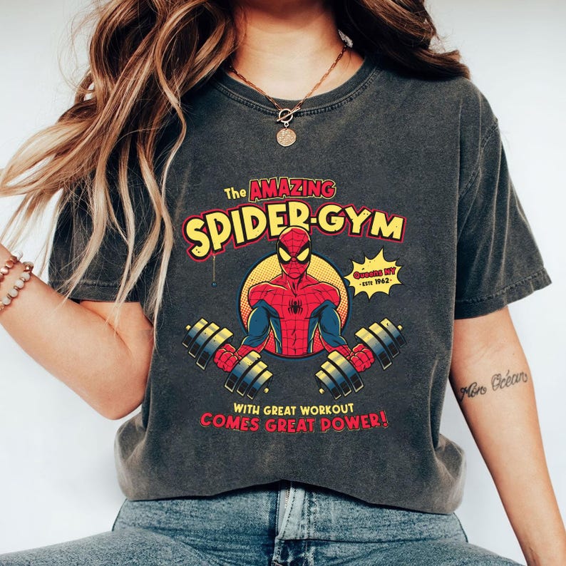 Może przedstawiać: Ciemnoszara koszulka z grafiką Spider-Mana podnoszącego ciężary, z napisem "The Amazing Spider-Gym". Projekt zawiera frazę "With great workout comes great power!" oraz ż&oacute;łto-czerwoną kolorystykę.