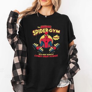 Może przedstawiać: Czarna koszulka z grafiką Spider-Mana. Projekt zawiera tekst "The Amazing Spider-Gym" i "With Great Workout Comes Great Power!" w kolorze czerwonym i ż&oacute;łtym. Grafika przedstawia Spider-Mana podnoszącego ciężary.