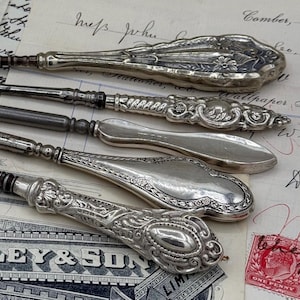 Peut inclure: Plusieurs outils anciens en argent, dont des crochets et autres instruments, sont disposés sur un fond de documents et d'éphémères anciens. Les outils présentent des manches ornés et des lames de différentes formes, pour une esthétique vintage.