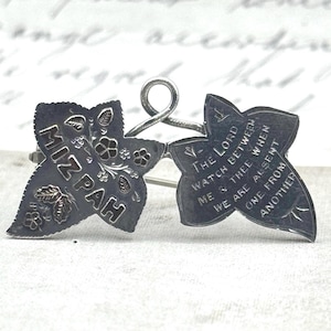 Op de afbeelding: Een zilverkleurige metalen broche in de vorm van twee klimopbladeren verbonden door een centrale lus. Het linkerblad bevat de gegraveerde tekst MIZPAH met bloemmotieven. Het rechterblad toont de gegraveerde tekst THE LORD WATCH BETWEEN ME & THEE WHEN WE ARE ABSENT ONE FROM ANOTHER.