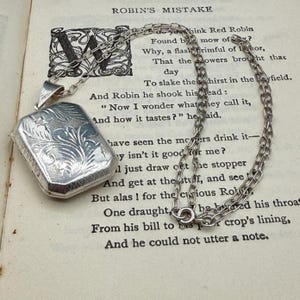 May include: A rectangular silver locket pendant featuring an intricate engraved floral pattern on the front surface, suspended from a matching silver link chain. The locket has a polished finish with decorative etched details along the edges.
