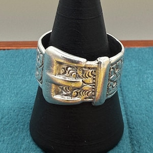 May include: A silver ring with a buckle design. The ring has an engraved floral pattern on the band and buckle. The buckle is rectangular with a detailed clasp. The ring is displayed on a black stand. The ring's design is reminiscent of a belt buckle.