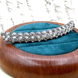 May include: A silver-toned, curved hair comb adorned with numerous clear, round gemstones. The comb is displayed on a teal velvet surface within a wooden jewelry box, showcasing its elegant design and sparkle.