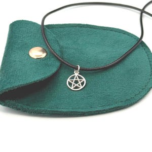 Puede incluir: Un colgante de pentagrama en tono plateado suspendido de un fino cordón negro. El colgante descansa sobre una pequeña bolsa de tela verde oscuro similar a la gamuza, con un botón de presión de metal dorado en la solapa superior.