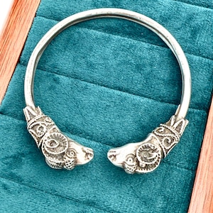 May include: A silver bracelet with ram head terminals. The open cuff bracelet is displayed on a teal velvet surface within a wooden frame. The ram heads feature detailed carvings.