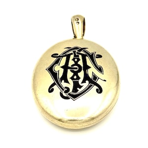 May include: A gold-colored, oval-shaped pendant with a black monogram design. The pendant has a small loop at the top for a chain. The monogram features intertwined letters, creating a decorative and elegant look. The pendant is likely made of metal.