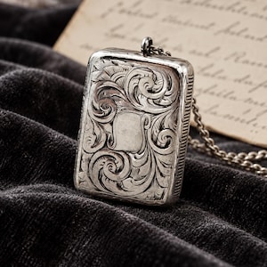 May include: A rectangular silver-toned metal locket pendant featuring an intricate engraved scrollwork pattern across the front surface. The pendant includes a small top loop attached to a matching silver-toned metal chain link necklace.