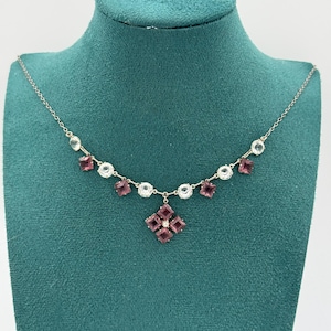 Op de afbeelding: Een delicate ketting met een zilveren ketting en een hanger met een cluster van vierkante, dieproze edelstenen. De ketting heeft ook heldere, ronde en vierkante edelstenen. De ketting wordt gepresenteerd op een groene fluwelen buste.