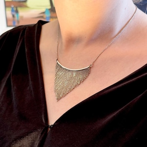 May include: A metallic gold-toned necklace featuring a curved bar at the top with a cascading, triangular fringe of small, interconnected metal beads that drape downward in a dense, textured pattern, suspended from a thin, delicate chain.