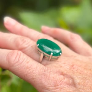 May include: A silver ring with a large, oval, polished green gemstone. The gemstone is set in a silver band with a simple design. The ring is displayed on a person's hand.