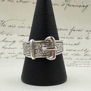 May include: A silver ring with a buckle design. The ring has a textured band and a detailed buckle clasp. The ring is displayed on a black cone-shaped stand against a backdrop of handwritten script.