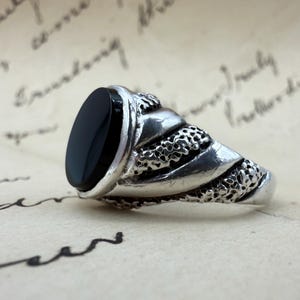 Può includere: Un anello d'argento con una pietra nera ovale. L'anello presenta un design dettagliato e strutturato attorno alla fascia. L'anello è posto su uno sfondo di carta invecchiata con scritte a mano.