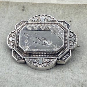 Pode incluir: Um broche retangular de prata com bordas arredondadas e ornamentadas, apresentando um painel central gravado. A gravação retrata um pássaro em voo sobre juncos, com uma borda de padrão geométrico. O metal tem um acabamento polido com detalhes oxidados.