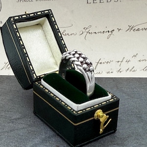 May include: A silver ring with a textured, clustered design is presented in a black and green jewellery box. The ring features small, rounded shapes. The box is open, revealing a green velvet interior and a gold-coloured clasp.