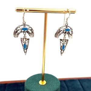 Puede incluir: Pendientes colgantes plateados con detalles en azul turquesa. Cada pendiente presenta una forma de media luna con detalles florales y un colgante triangular. Los pendientes se exhiben sobre un soporte dorado sobre un fondo blanco.