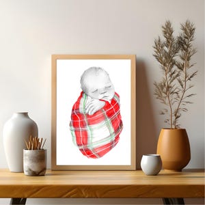 May include: A framed watercolor print of a sleeping baby wrapped in a red and green plaid blanket. The artwork is in a natural wood frame and rests on a wooden surface. Decorative vases and a plant are in the background.
