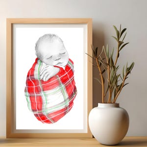 May include: A framed watercolor print of a sleeping baby wrapped in a red, green, and white plaid blanket. The artwork is displayed on a wooden surface, with a white vase and greenery in the background.