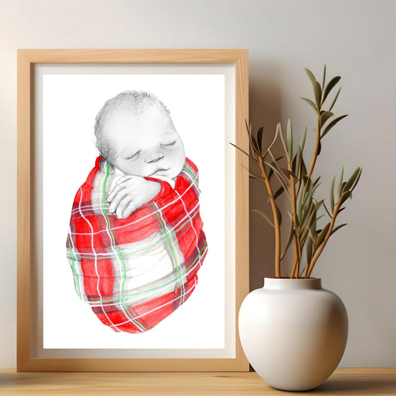May include: A framed watercolor print of a sleeping baby wrapped in a red, green, and white plaid blanket. The artwork is displayed on a wooden surface, with a white vase and greenery in the background.